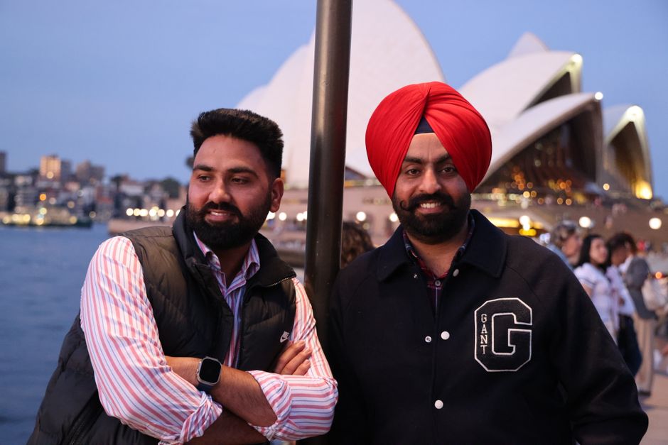 Two men of South Asian descent stand outdoors near the Sydney Opera House at dusk, with the water and city skyline visible in the background. The man on the left has short dark hair, a full beard, and