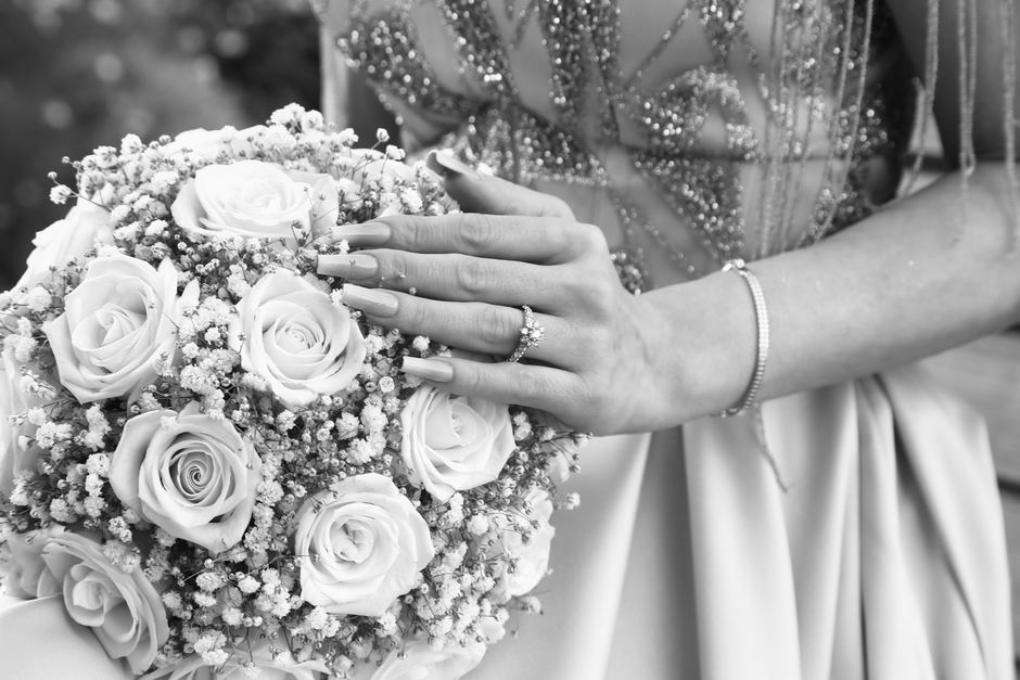 A close-up black and white photograph showing a woman in an elegant, embellished dress holding a large, round bridal bouquet of fresh roses and baby's breath. Her left hand rests on the bouquet, displ