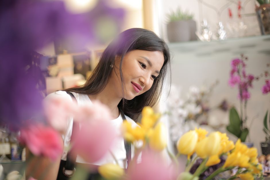 A young woman with long dark hair, wearing a white top, is in a florist shop dedicated to flower delivery and arrangements for Gants Hill offices and shops. She is carefully observing a bouquet of fre