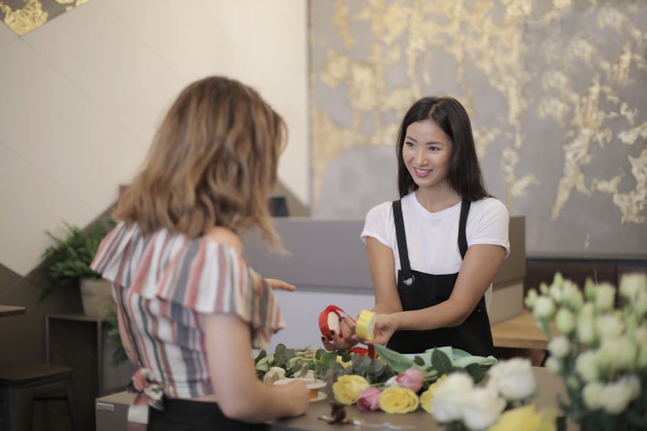 A professional florist from Gants Hill Florist, wearing a white t-shirt and black apron, smiling as she hands a small, decorated floral gift to a customer. The customer, with shoulder-length blonde ha