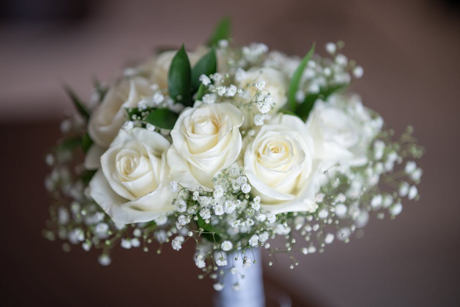A bridal bouquet featuring ivory roses with lush, velvety petals, accented by sprigs of delicate white baby's breath and green foliage. The bouquet is arranged in a rounded, compact style and wrapped 